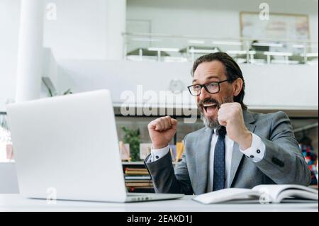 Excited joyful middle aged businessman sits at the desk, overjoyed with financial results. Happy caucasian male employee well done with project or got promotion on work, success concept Stock Photo