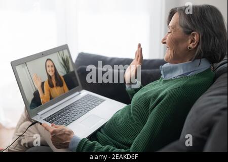 A middle-aged gray-haired lady wearing a green sweater jumper is looking and waving at the laptop screen, grandmother senior lady is talking to her Stock Photo