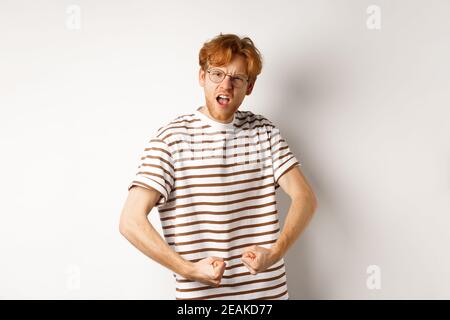 Image of confident and strong redhead man flexing biceps, showing muscles after gym, standing over white background Stock Photo