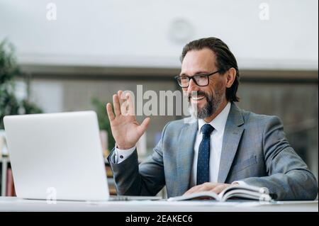 Friendly successful businessman sitting in modern office, using laptop, having online conference. Smiling male employee on video briefing meeting, discussing work process with colleagues Stock Photo