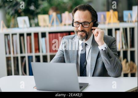 Caucasian male employee in headphones uses laptop, sitting at the desk. Senior satisfied businessman is consulting clients, smiling, work in support service as call center operator Stock Photo