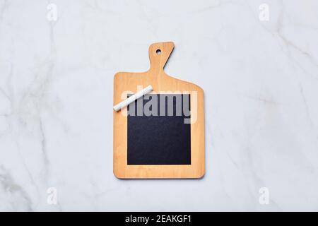 Chalk board in the form of kitchen cutting board with piece of chalk on light marble table. Board for writing notes, lists, recipes Stock Photo