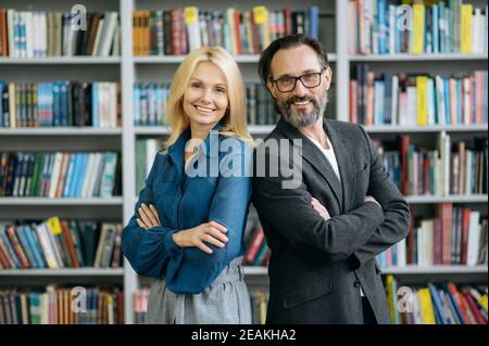 Portrait of mature successful business partners standing with arms crossed in modern office. Elegant male and female colleagues in formal wear look directly at the camera, smiling, teamwork concept Stock Photo