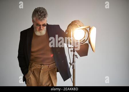 Portrait of classy bearded middle aged man dressed elegantly standing next to studio spotlight while posing for camera over white background. Fashion photoshoot, style concept Stock Photo