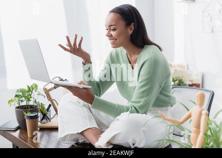 cheerful african american freelancer sitting on desk and gesturing during video chat on laptop, blurred foreground Stock Photo