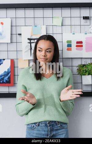 young african american interior designer gesturing while standing near paintings on wall Stock Photo