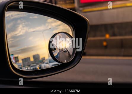 Reflection of traffic flow on asphalt road in side mirror of blue SUV. Car wing mirror with convex mirror for safety driving. View on the road and sunset sky behind cityscape through car side mirror. Stock Photo