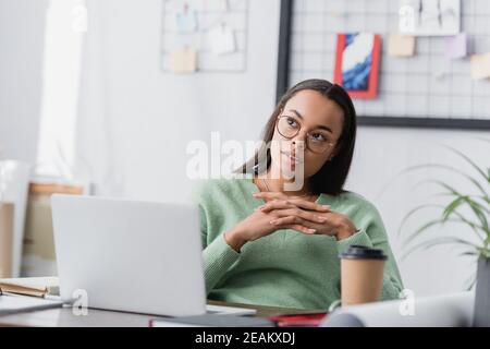 thoughtful african american architect looking away while sitting near laptop at home studio, blurred foreground Stock Photo