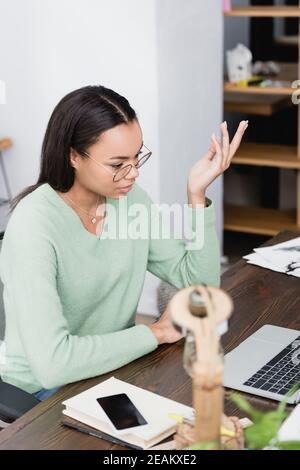 african american interior designer in eyeglasses gesturing near laptop on blurred foreground Stock Photo