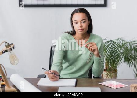 pensive african american interior designer holding pencil and eyeglasses while sitting near sketchbook on desk, blurred foreground Stock Photo