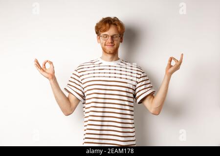 Calm and relaxed young man with red messy hair, spread hands sideways in mudra gesture and smiling, practice yoga or meditating, white background Stock Photo