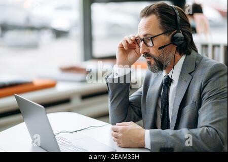 Focused male operator of call center or ceo is sitting at the desk, having a serious conversation. Senior business man is wearing eyeglasses, formal suit and headset communicate online Stock Photo