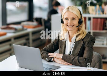 Joyful elegant female operator of call center in headset is looking at the camera, smiling. Intelligent business woman is sitting at the table in office, working in support service, consulting people Stock Photo