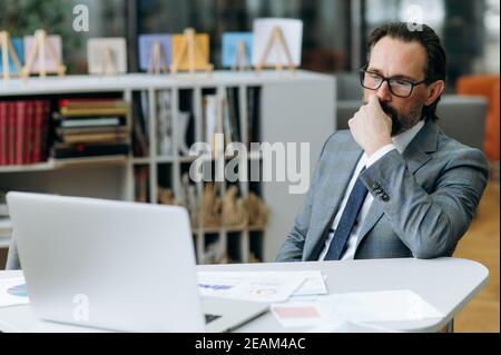 Middle aged businessman dressed in elegant suit, concentrated on serious project, sitting at the desk in modern office. Focused male manager is using laptop, thinking about business strategy Stock Photo