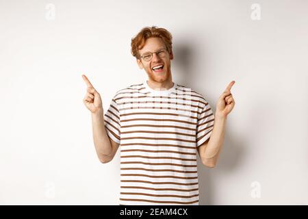 Happy hipster guy with red messy hair and glasses smiling, pointing fingers up, showing sale promotion, white background Stock Photo