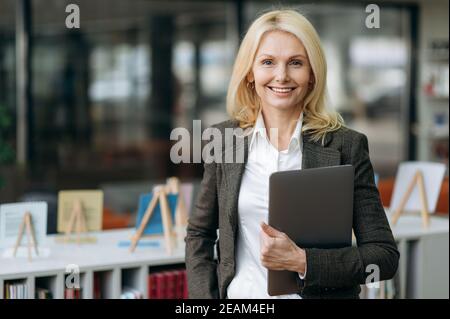 Portrait of influential elegant businesswoman with blonde hair in stylish formal suit. Happy successful middle aged lady looks directly at the camera, friendly smiling, holding laptop in arms Stock Photo
