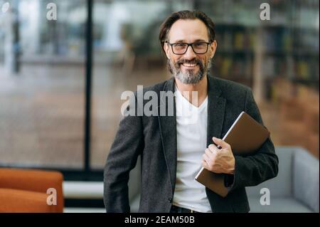Portrait of happy confident businessman in eyaglasses and stylish formal suit. Middle aged successful male business leader looking directly at the camera, smiling, holding laptop in arms Stock Photo