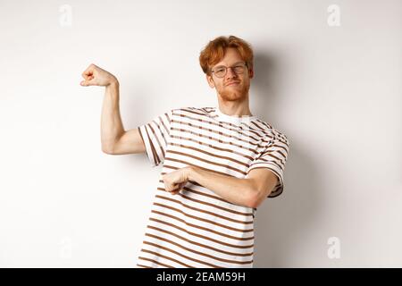 Image of confident and strong redhead man flexing biceps, showing muscles after gym, standing over white background Stock Photo