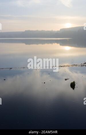 Vertical hazy sunset reflected in calm water with edge of reservoir Stock Photo