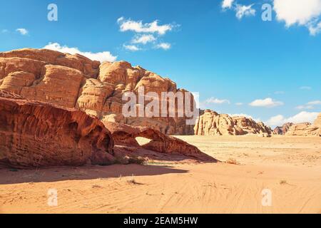 Rocky massifs on red sand desert, small stone arc bridge, bright blue sky in background - typical scenery in Wadi Rum, Jordan