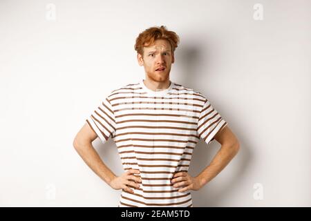 Confused and offended man with ginger curly hair staring at camera puzzled, standing questioned against white background Stock Photo