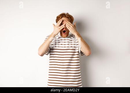 Shocked and embarrassed redhead guy covering eyes, peeking through fingers and gasping, standing over white background Stock Photo