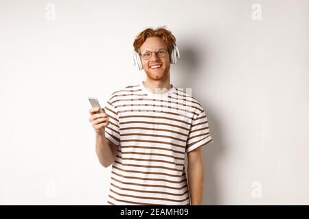 Technology concept. Young man with red hair and beard listening music in headphones and using smartphone, smiling at camera, white background Stock Photo