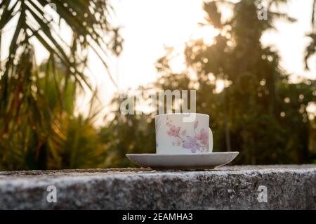 Coffee cup in sunset sunlight. Summer fresh cool look. White coffee cup on saucer for hot drink on roof beam of a residential building with bokeh city in the background. Stock Photo