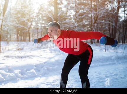 Outdoor winter strength training. Positive senior sportsman with dumbbells doing powerlifting exercises in snowy park Stock Photo