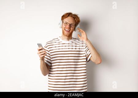 Technology concept. Young man with red hair and beard listening music in headphones and using smartphone, smiling at camera, white background Stock Photo