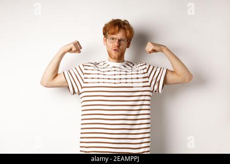 Image of confident and strong redhead man flexing biceps, showing muscles after gym, standing over white background Stock Photo