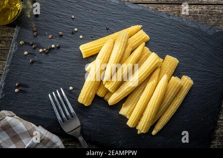 Pickled young baby corn cobs on black stone cutting board. Stock Photo