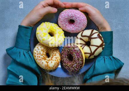 Two hands holding plate of colourful sugary round glazed donuts with colored sprinkles on gray background top view Stock Photo