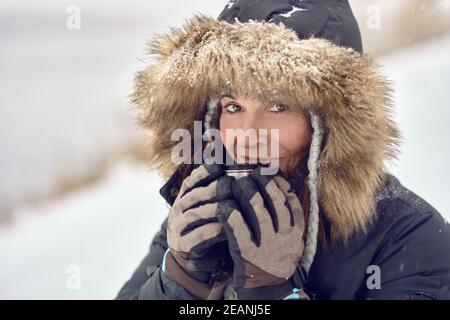 Happy smiling woman wearing a fur trimmed hooded jacket enjoying a mug of coffee cradled in her gloved hands outdoors in winter snow in a concept of t Stock Photo