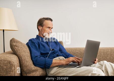 Side view of senior man wearing earphone typing on laptop. Elderly male sitting on sofa with laptop  Stock Photo