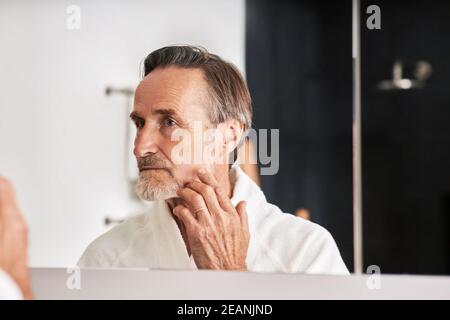 Senior man touching her face in bathroom in from of a mirror. Male in bathrobe looking at his reflection  Stock Photo