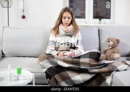 Cold and flu season. Sick teenage girl with high fever and sore throat, holding cup of hot tea and feeling bad, sitting at home on the sofa under warm Stock Photo