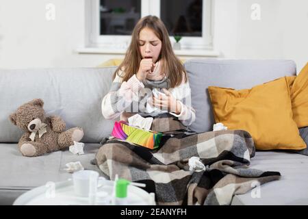 Pleasant tired sick teen girl with scarf around neck, covered with blanket, sitting on soft couch and coughing, suffering from flu or cold and sore Stock Photo