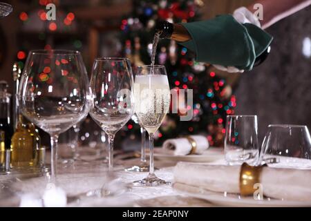 Champagne is poured into a glass on the background of a Christmas tree. Sparkling wine. Unrecognizable waiter. Festive table setting. Stock Photo