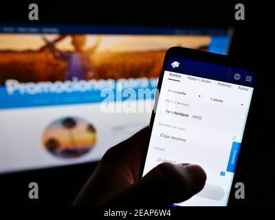 Person booking flight on website of airline Aerovías de México (Aeroméxico) on smartphone in front of screen. Focus on top of cell phone display. Stock Photo