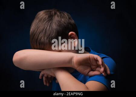 crying european teenage boy with light brown hair, wearing blue sports t-shirt, covering his face with hand over isolated background Stock Photo