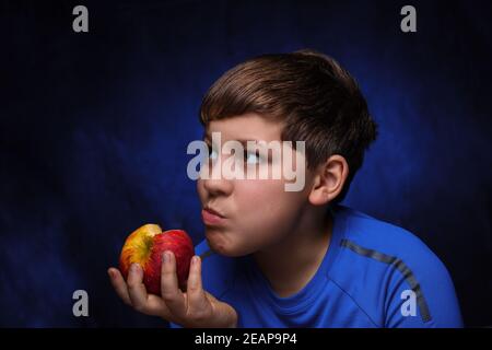 big red-yellow apple in the hand of eating it European teenage boy with light brown hair, dressed in a blue sports t-shirt, on an isolated background Stock Photo
