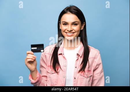 Beautiful caucasian brunette girl in stylish clothes holds a bank credit card in her hand, looks and smiles at the camera, standing on an isolated blue background Stock Photo