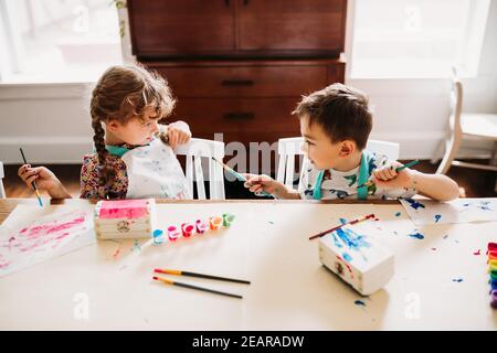 Brother and sister sitting at table painting with rainbow paints Stock Photo