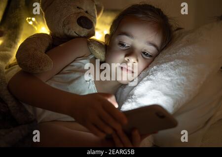 A cute little girl lies on a pillow with a teddy bear and plays on the phone at night in bed. Stock Photo