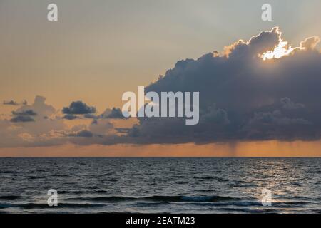 Sun shining through dark cumulus clouds sky above sea surface in evening. Fluffy stormy cloudscape with sunlight over dark blue sea Stock Photo