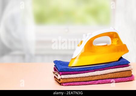 Closeup of a yellow iron on a stack colorful towels on table against an abstract bright curtain backgound. Advertising for the laundry service with space for design. Stock Photo