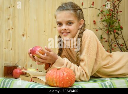 A girl lying down reads a book, holds an apple in her hands and looks happily into the frame Stock Photo