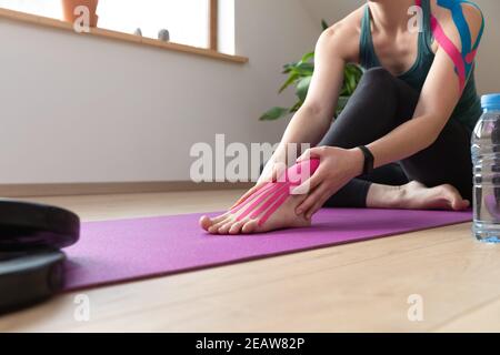Young beautiful women showing on her foot applied elastic kinetic tape on a sunny day in attic. Workout at home. Kinesiology physical therapy. Stock Photo