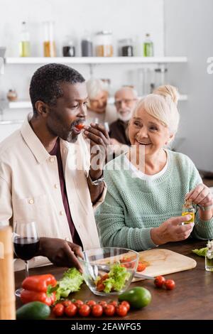 African american man eating cherry tomato near senior woman and retired friends on blurred background Stock Photo